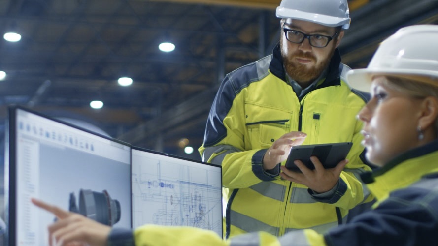 A man and a woman in hard hats and high visibility jackets looking at two computer screens with images of computer models on them.