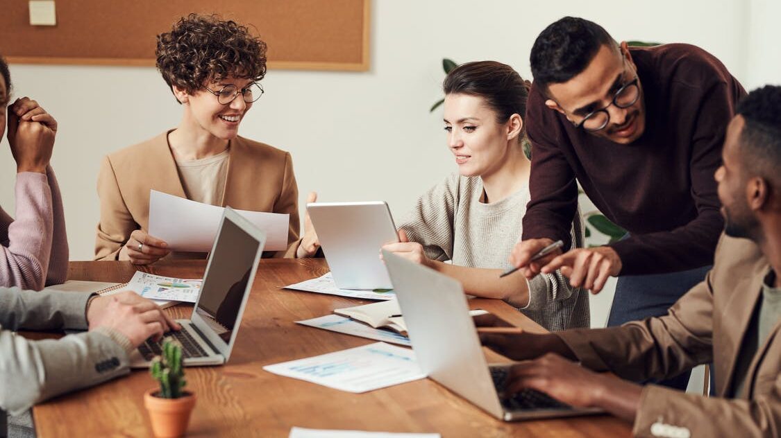 A group of employees sit with laptops around a meeting table.