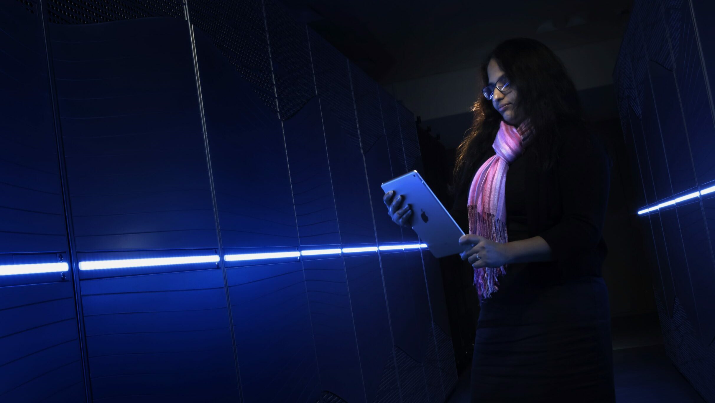 Woman holding a tablet device standing in front of a supercomputer illuminated in blue light.