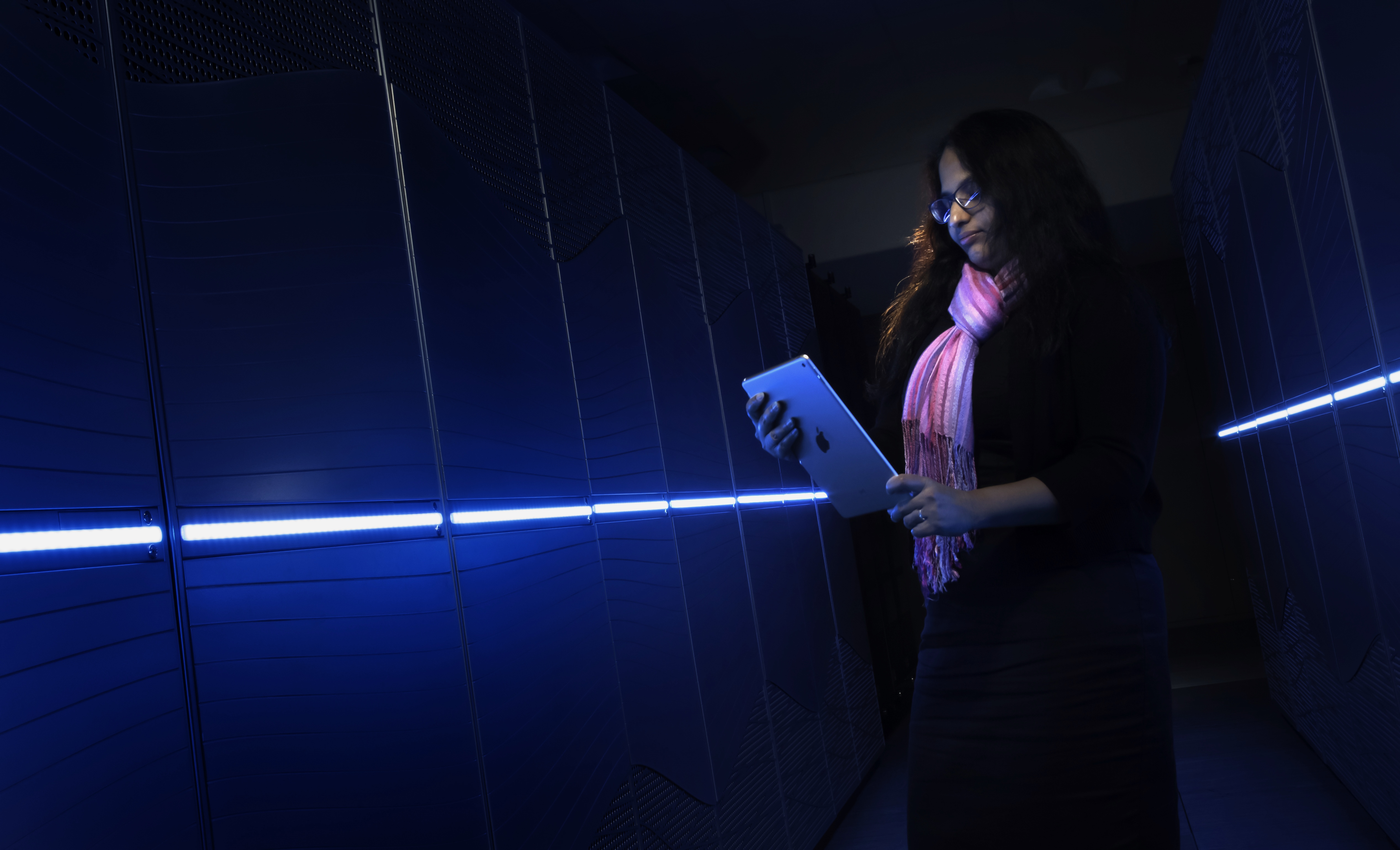 Woman holding a tablet device standing in front of a supercomputer illuminated in blue light.