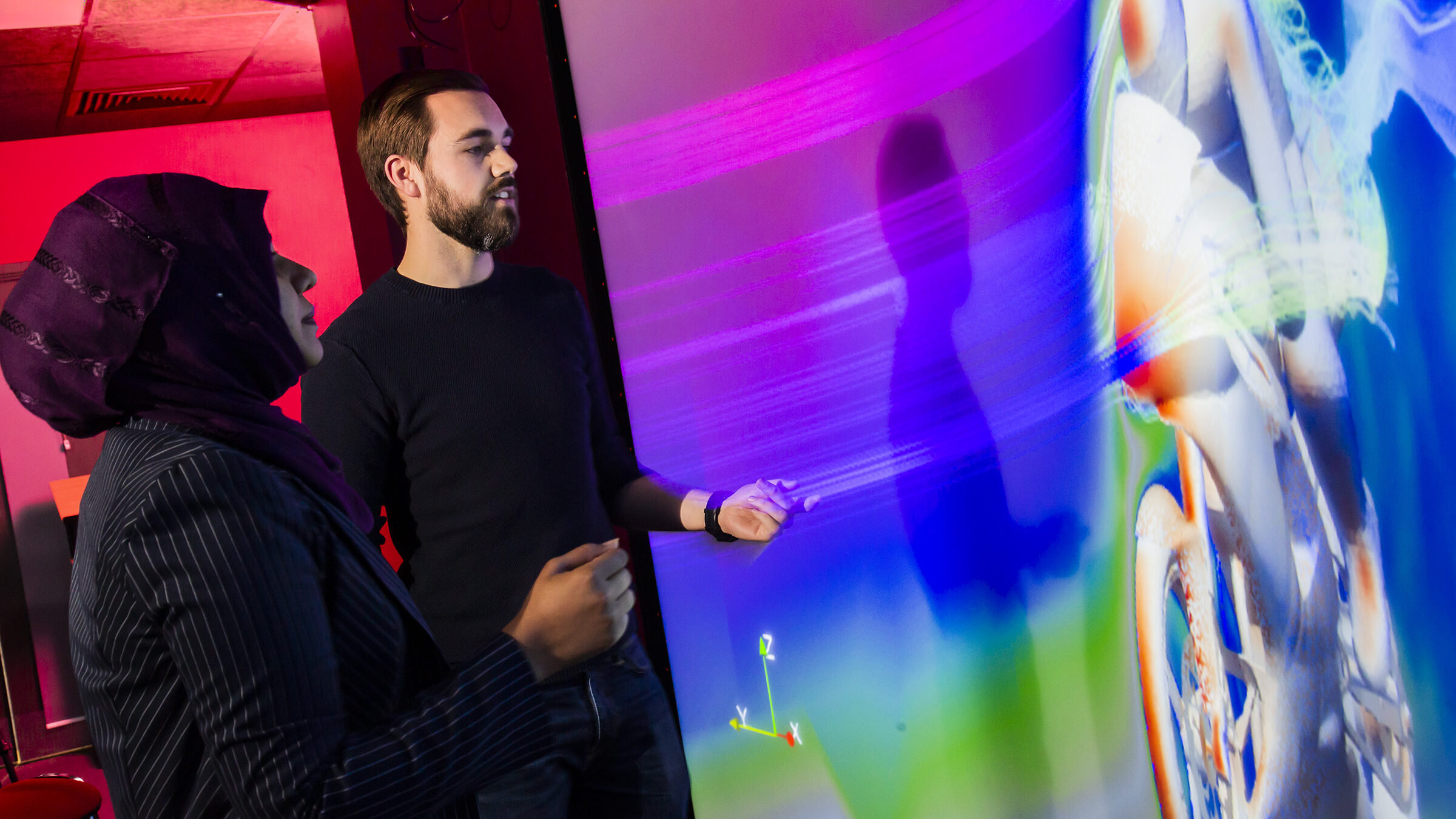 A man and woman standing in front of a visualisation screen bigger than they are, looking at streamlines over a motorbike simulation.