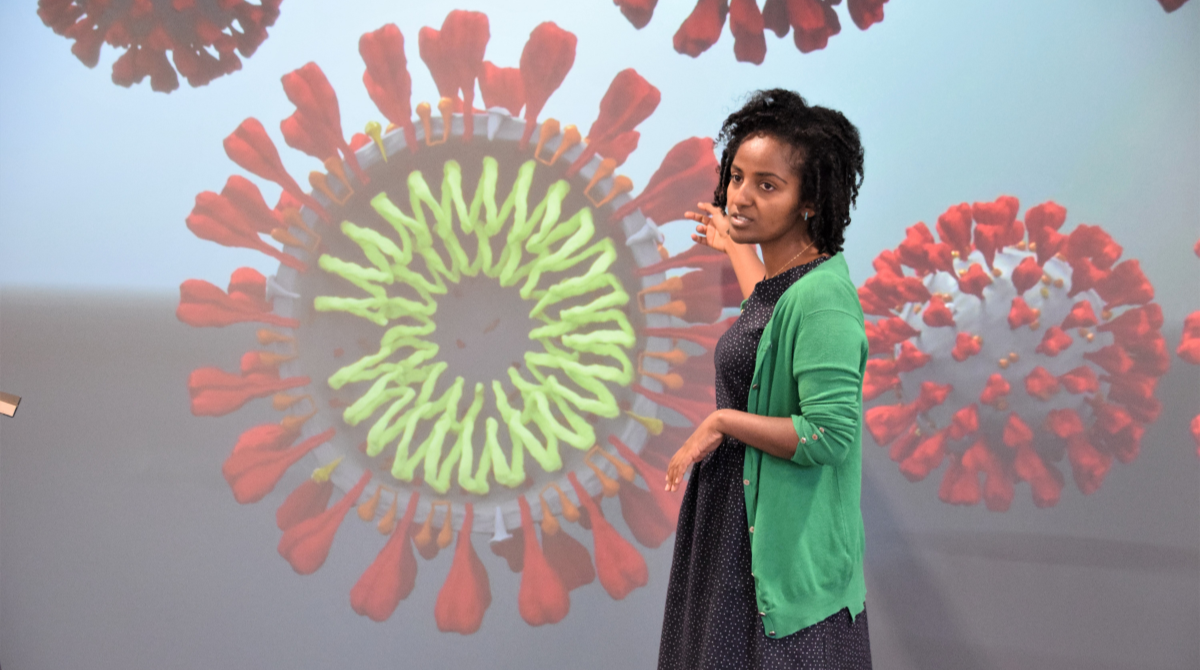 Woman standing in front of a visualisation screen gesturing at a simulation of disease molecules