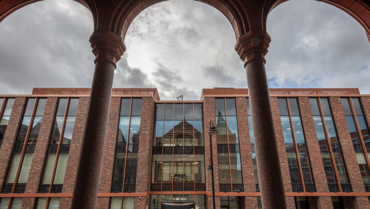 The Digital Innovation Facility, a modern red drick building with many windows, viewed through a more traditional historic archway of an older university building opposite it.