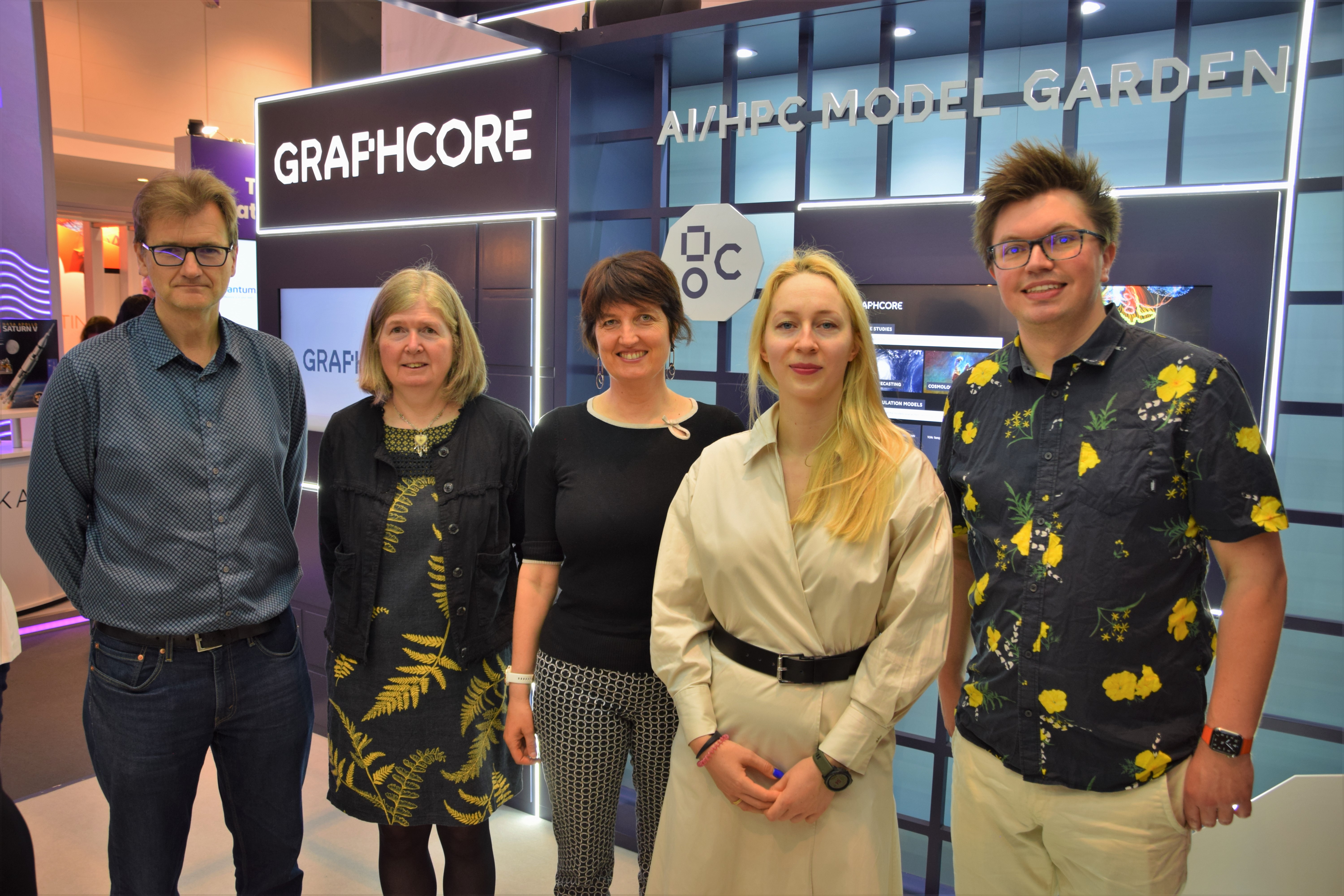 A group of two men and three women stand in front of the Graphcore exhibition stand at a conference.