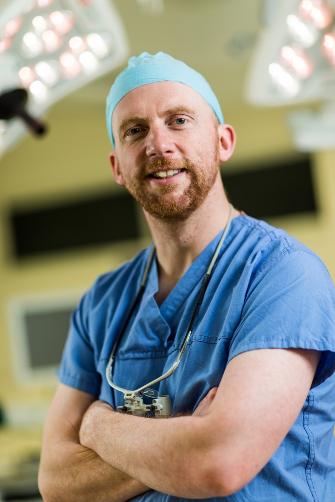 Headshot of Iain Hennessey wearing scrubs, arms folded with a stethoscope around his neck.