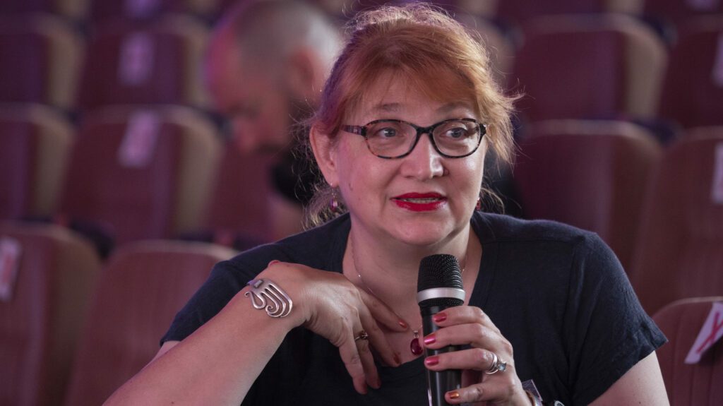 A woman holding a microphone sitting among a conference audience.