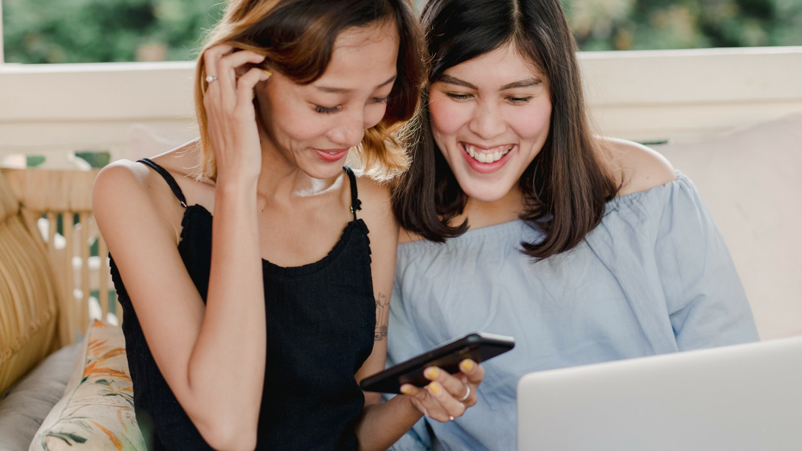 Women smiling sitting next to each other with a laptop in front of them and looking at a phone in their hands.