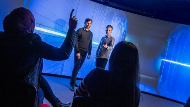 Two presenters stand in front of a curve visualisation wall. Two audience members are visible in silhouette. One has their hand up.