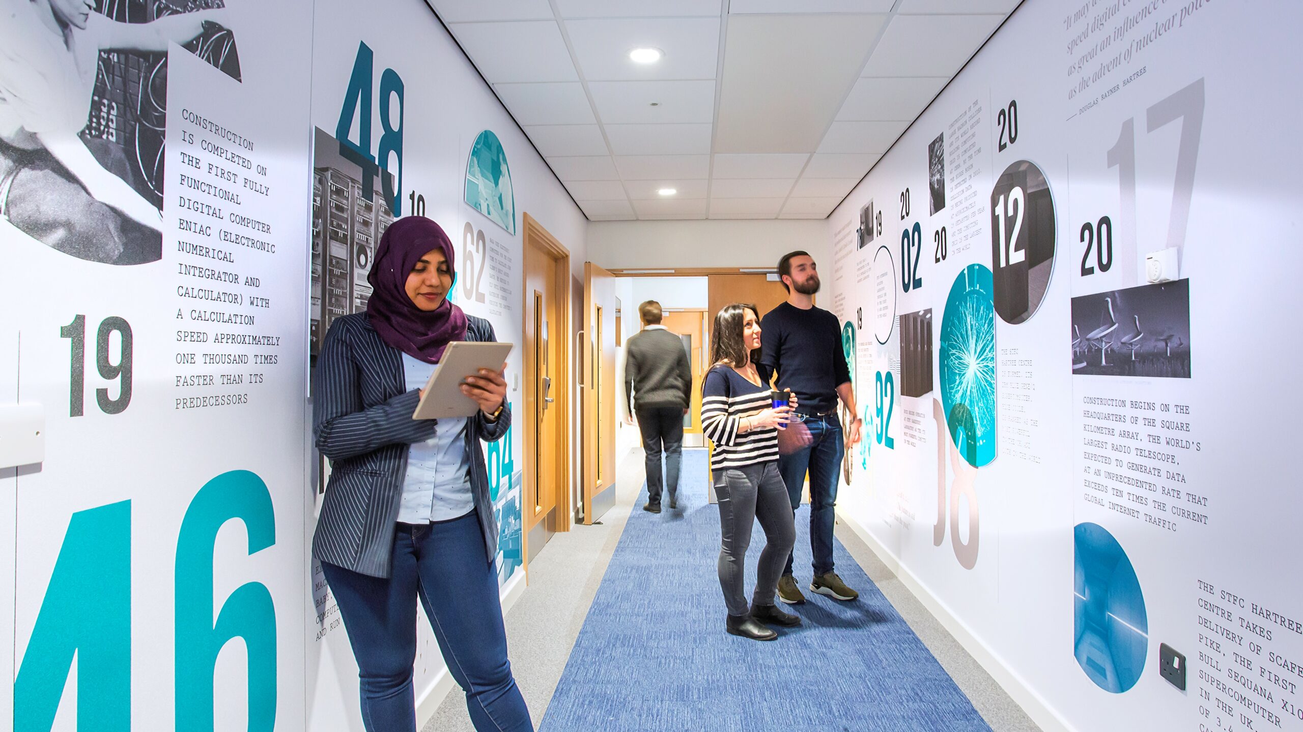 People standing in a corridor chatting. One woman is looking at a tablet device in her hands.