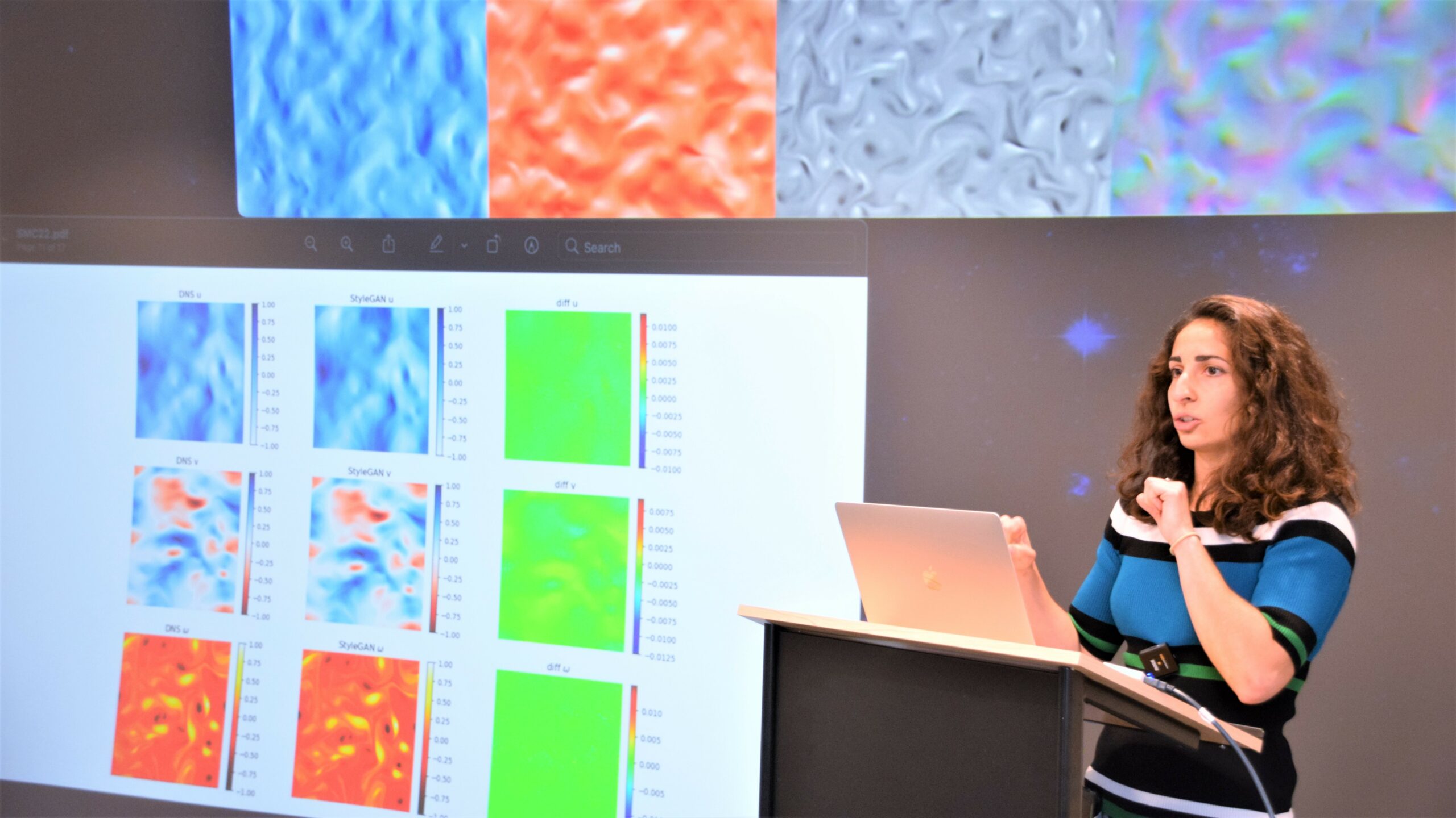 A woman stands at a lectern in front of a large screen displaying colourful simulations and scientific results.