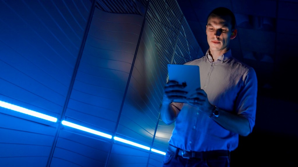 A man stands in front of a supercomputer, looking at a tablet device.