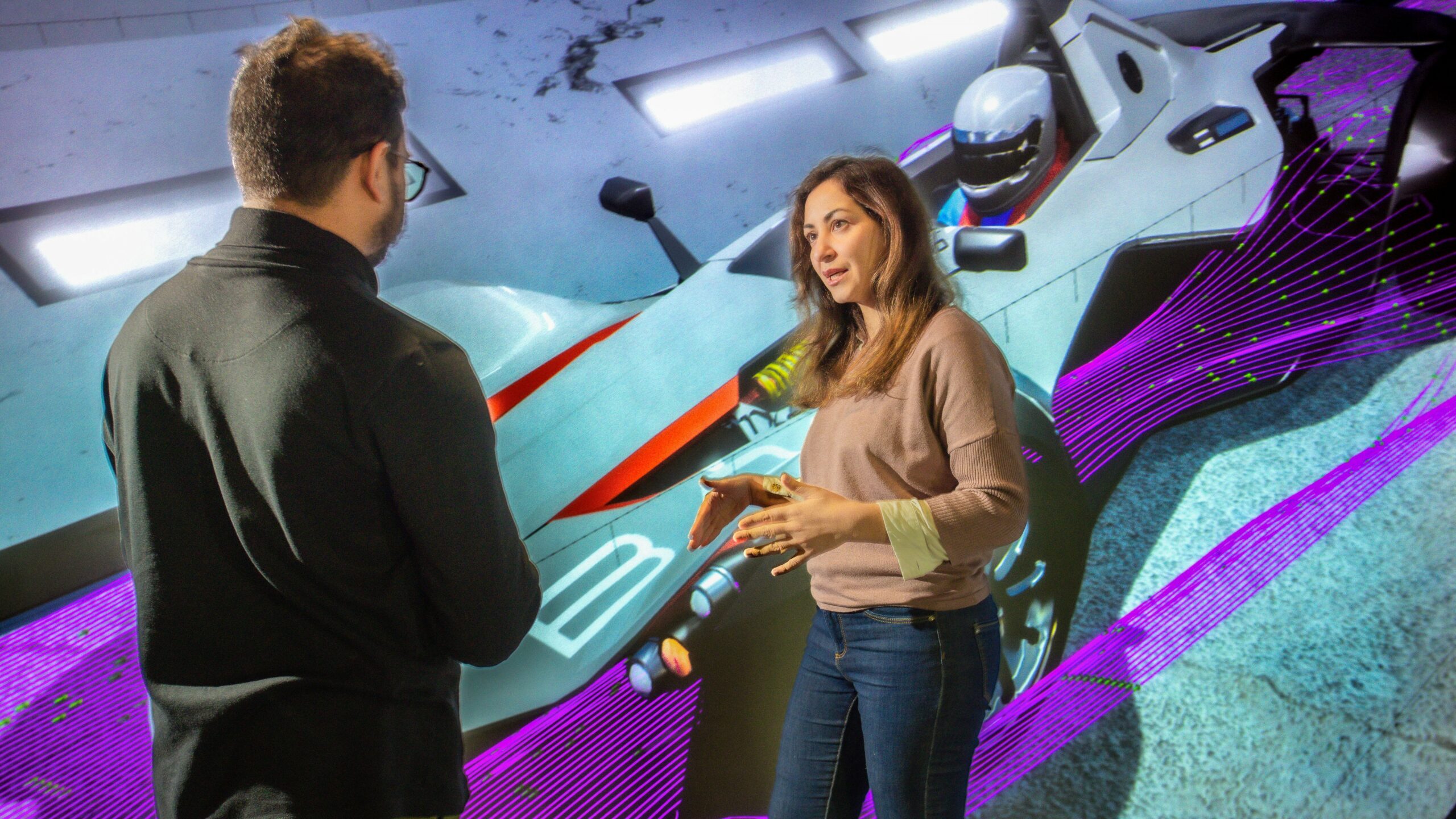 A woman and a man stand in front of a virtual wind tunnel visualisation showing streamlines over a racecar.