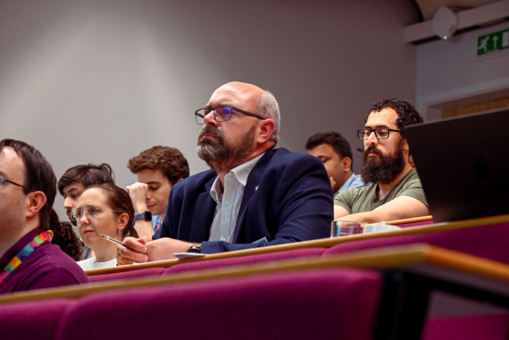 A white man in a suit listening to a talk in a crowded lecture theatre.