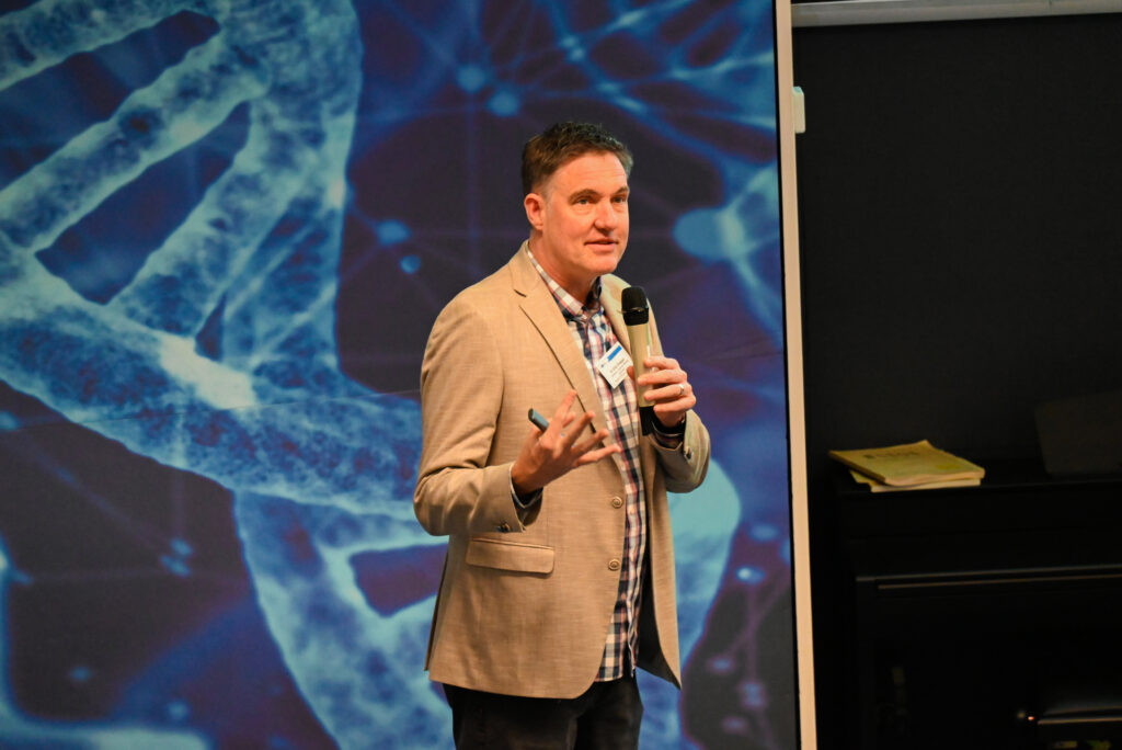 A white man in a blazer holding a microphone speaking to attendees in a lecture theatre.