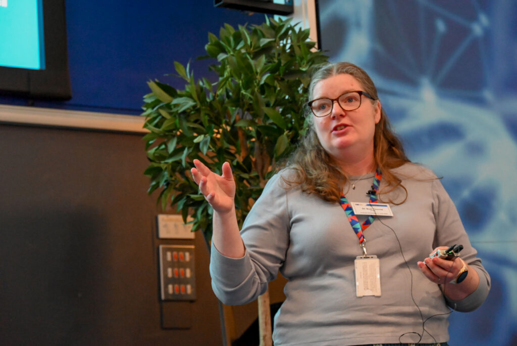 A white woman in glasses, holding a microphone speaking to an audience in a lecture theatre.